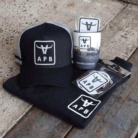 A black and gray APB trucker hat displayed with matching merchandise, including a black t-shirt, a sticker, and a branded cup, arranged on a textured concrete surface.