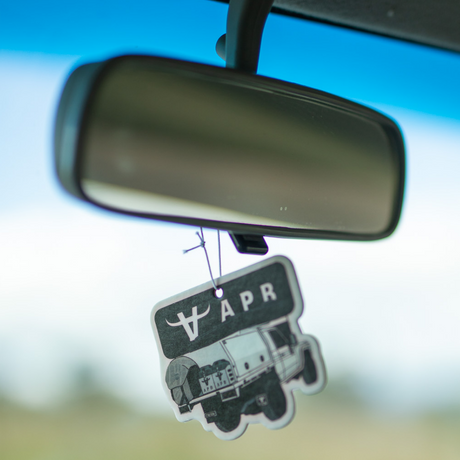 A black and white air freshener or car fragrance tag shaped like a military vehicle, hanging from a rearview mirror inside a car with a blurred blue sky and outdoor background.