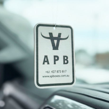 A white and black APB car air freshener hanging from the windshield of a vehicle, showing the brand logo and contact details.